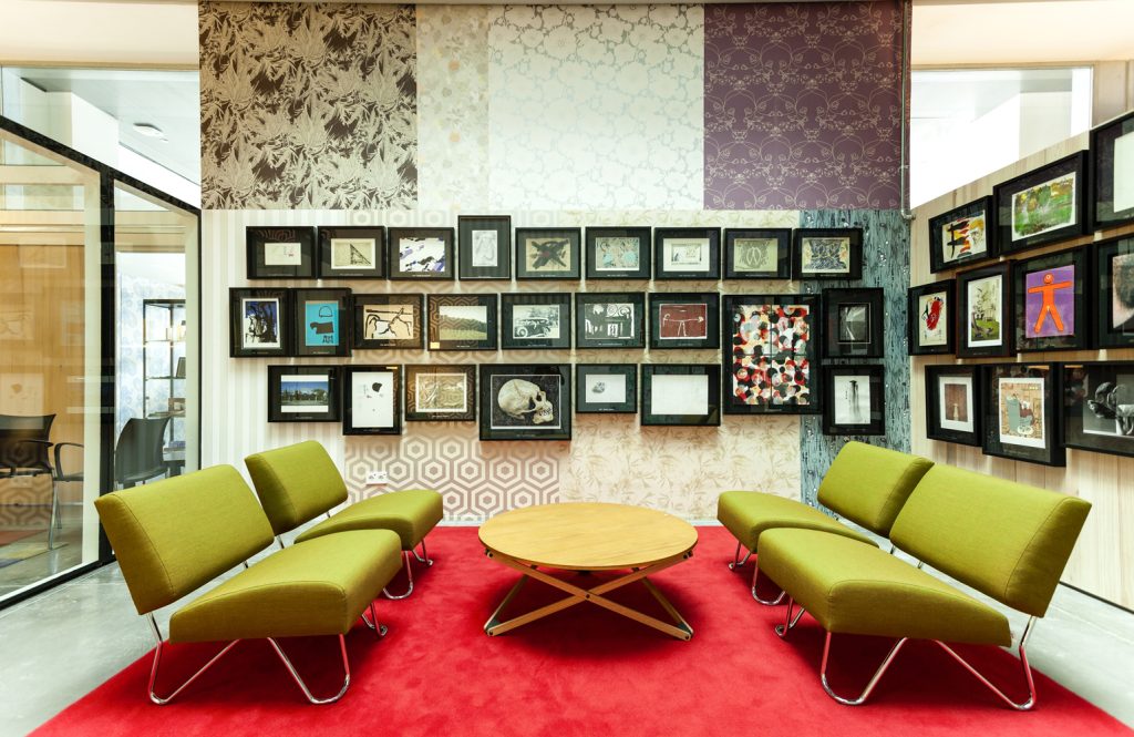DHUB offices in Barcelona - BAAS Architecture - Modern, artistic lounge area with green chairs, a wooden round table, and a wall adorned with various framed artworks