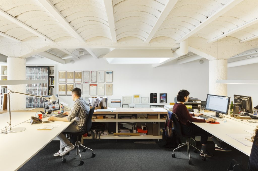 BAAS Studio - BAAS Arquitectura - Two individuals collaborate at a shared desk in a bright, industrious office setting