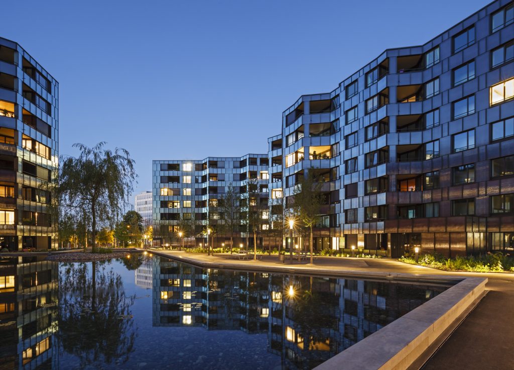 Guggach Residential Building - BS + EMI Architektenpartner AG - Serene evening view of modern, illuminated apartment buildings reflecting in a calm fountain