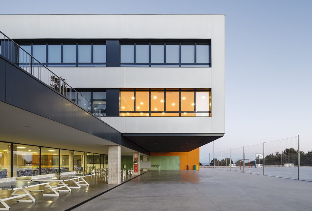 Hamelin School - Alonso i Balaguer - Modern building with a protruding upper level illuminated from within, overlooking an outdoor area with benches and a sports court