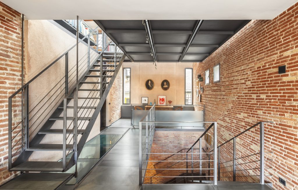 Savona Flat - Guido Canali - Modern, spacious interior with an industrial aesthetic, featuring a metal staircase, glass flooring, and exposed brick walls.