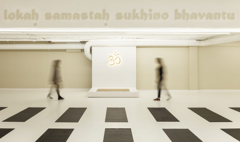 YogaOne DIR - Studio Shito - Blurred figures move through a yoga-themed space, under the mantra “lokah samastah sukhino bhavantu” and an Om emblem